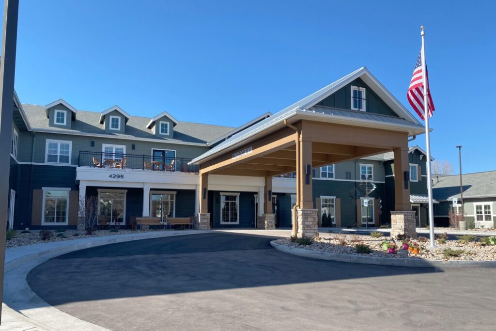 Two-story building with green siding, white trim, and a large covered drive-up entrance. An American flag stands at the front, and the landscaping includes flowers and stone beds. Balconies and dormer windows add to the residential-style design.