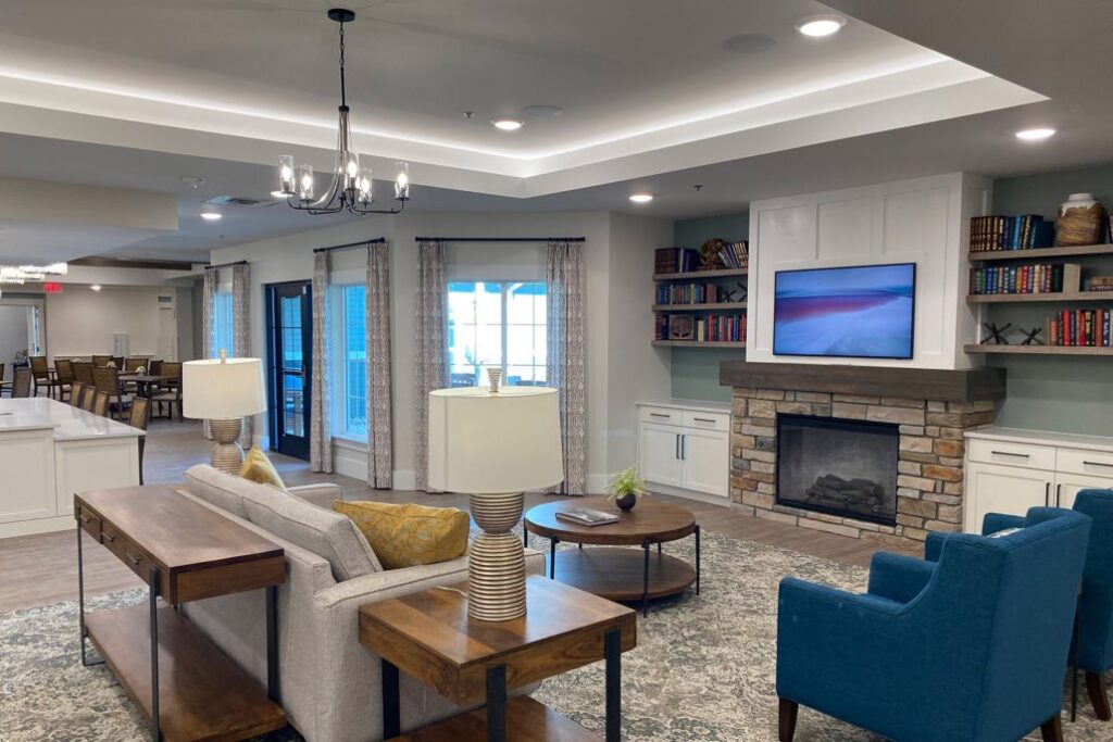 Inviting lounge with a stone fireplace, built-in bookshelves, and a mounted TV. The space features a sofa, blue armchairs, wooden tables, and warm lighting, with dining tables visible in the background.