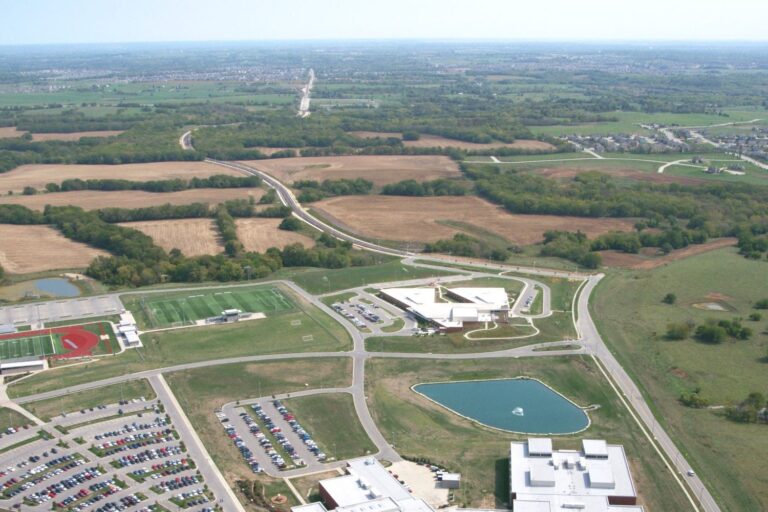 Curving roadway runs through open fields, past a school with parking lots, sports fields, and a pond, connecting to distant neighborhoods.