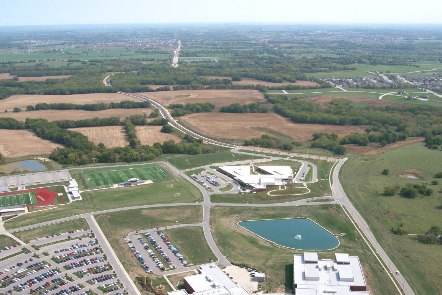 Curving roadway runs through open fields, past a school with parking lots, sports fields, and a pond, connecting to distant neighborhoods.