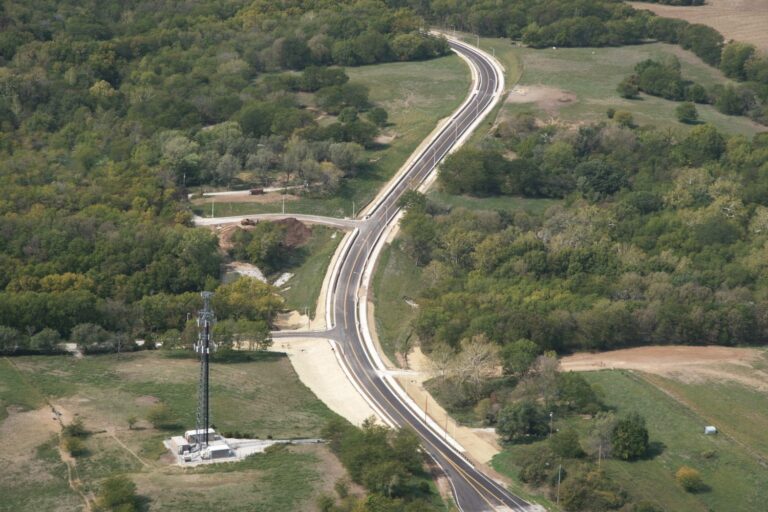 Freshly paved road with bright lane markings winds through a rural area with dense trees, fields, and a tall communication tower nearby.