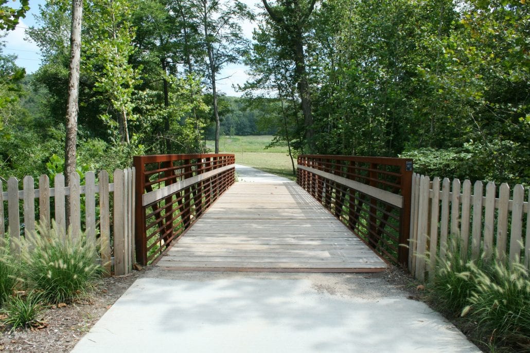 Wooden walkway bridge with rust-colored metal railings and picket fences on both sides, leading through a wooded area toward an open grassy field under a clear sky.