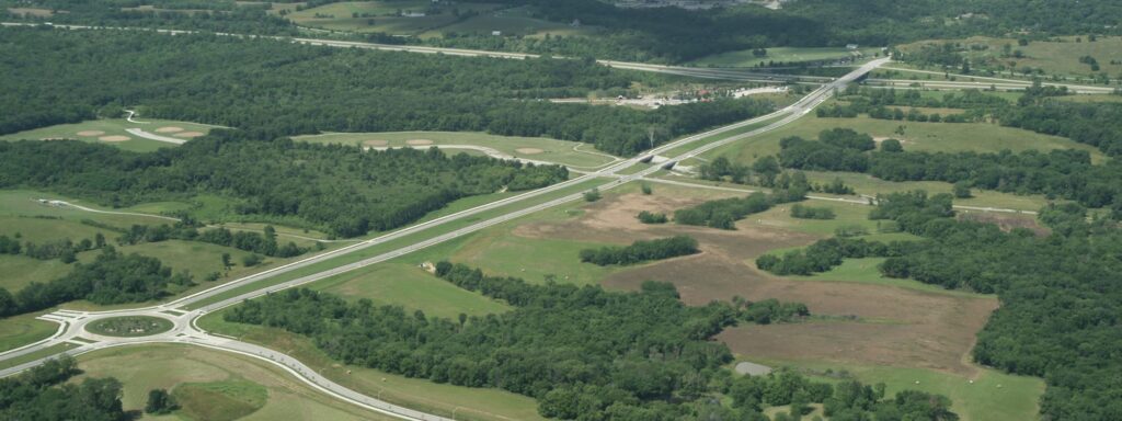 Expansive aerial scene showing a curving parkway with roundabouts cutting through lush green fields, wooded areas, and open meadows under a clear sky.
