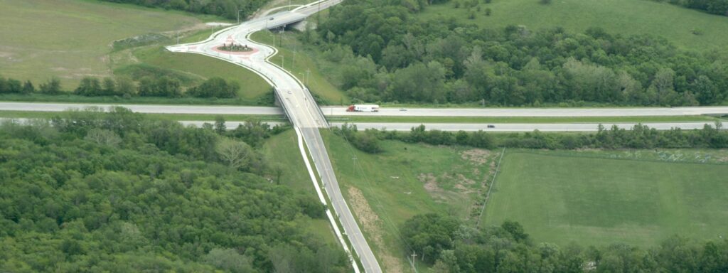 Elevated highway interchange showing a curved roadway crossing over a main highway, bordered by dense green trees and open grassy fields.