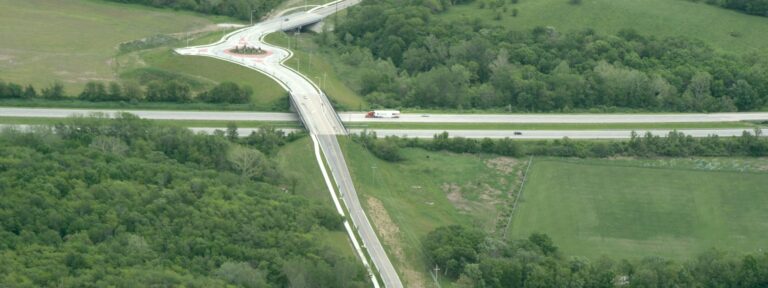 Elevated highway interchange showing a curved roadway crossing over a main highway, bordered by dense green trees and open grassy fields.