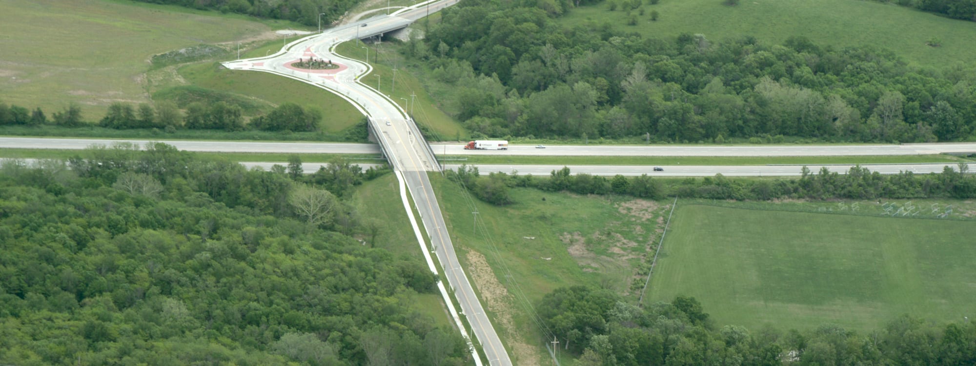 Elevated highway interchange showing a curved roadway crossing over a main highway, bordered by dense green trees and open grassy fields.