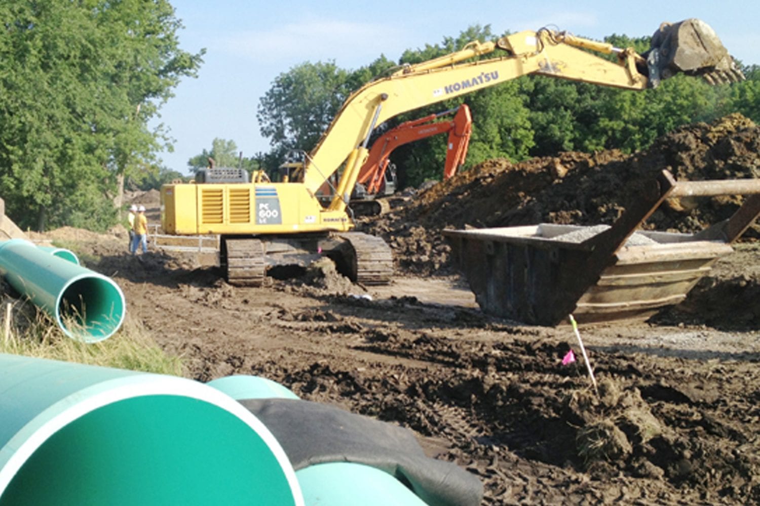 Large yellow excavator digging on a muddy construction site with green PVC pipes in the foreground, workers nearby, and piles of dirt surrounded by trees.