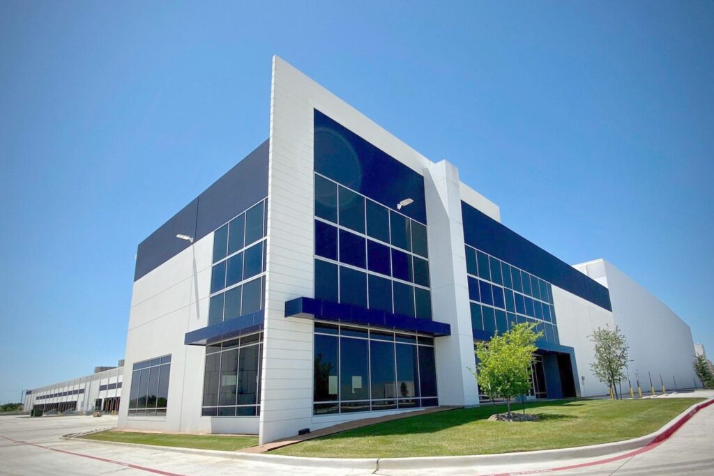 Large contemporary warehouse featuring reflective blue-tinted windows, white metal exterior, and landscaped surroundings under a bright blue sky.