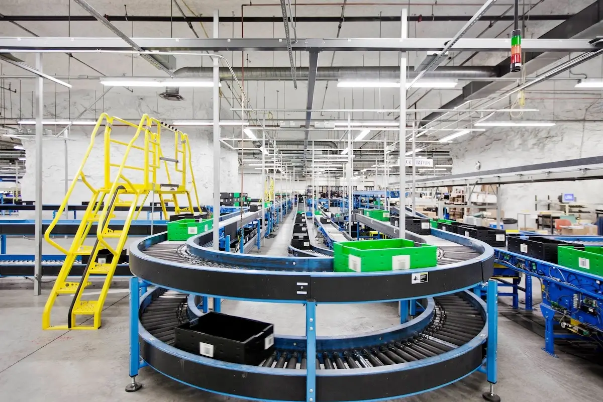 Curved conveyor belts inside a warehouse move green and black plastic bins, with a yellow safety ladder nearby.