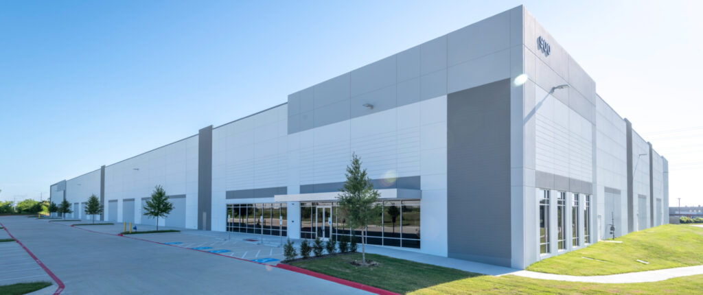 Large contemporary warehouse with white and gray exterior panels, reflective glass entryways, and small landscaped trees along a wide concrete driveway under a bright blue sky.