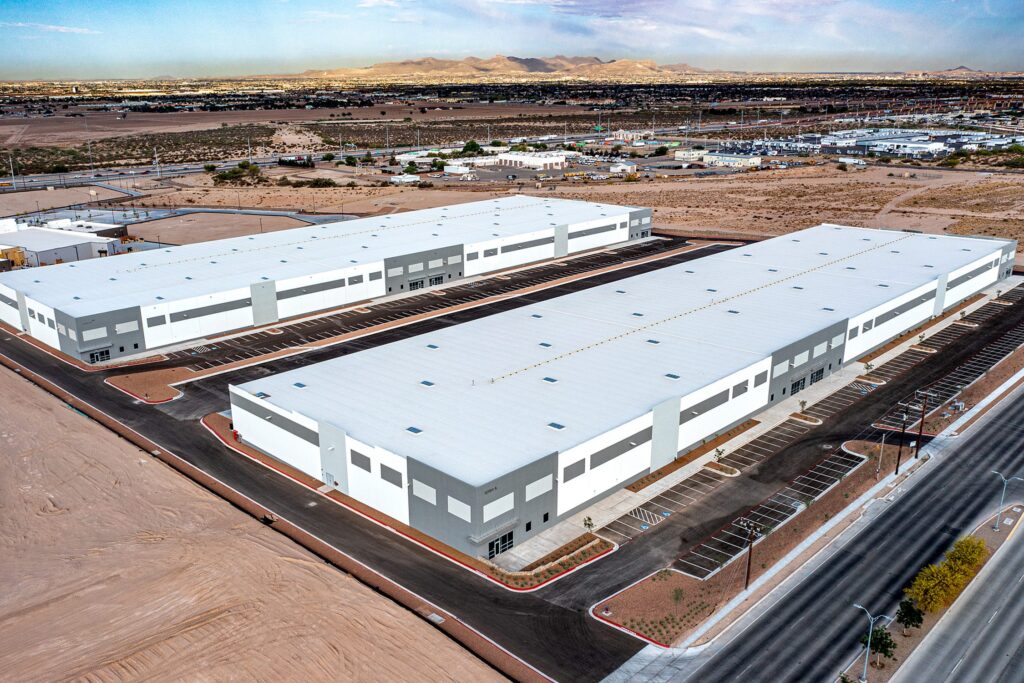 Two large, white and gray industrial warehouse buildings with flat roofs and adjacent parking lots in a desert landscape, surrounded by roads and distant mountains under a clear sky.