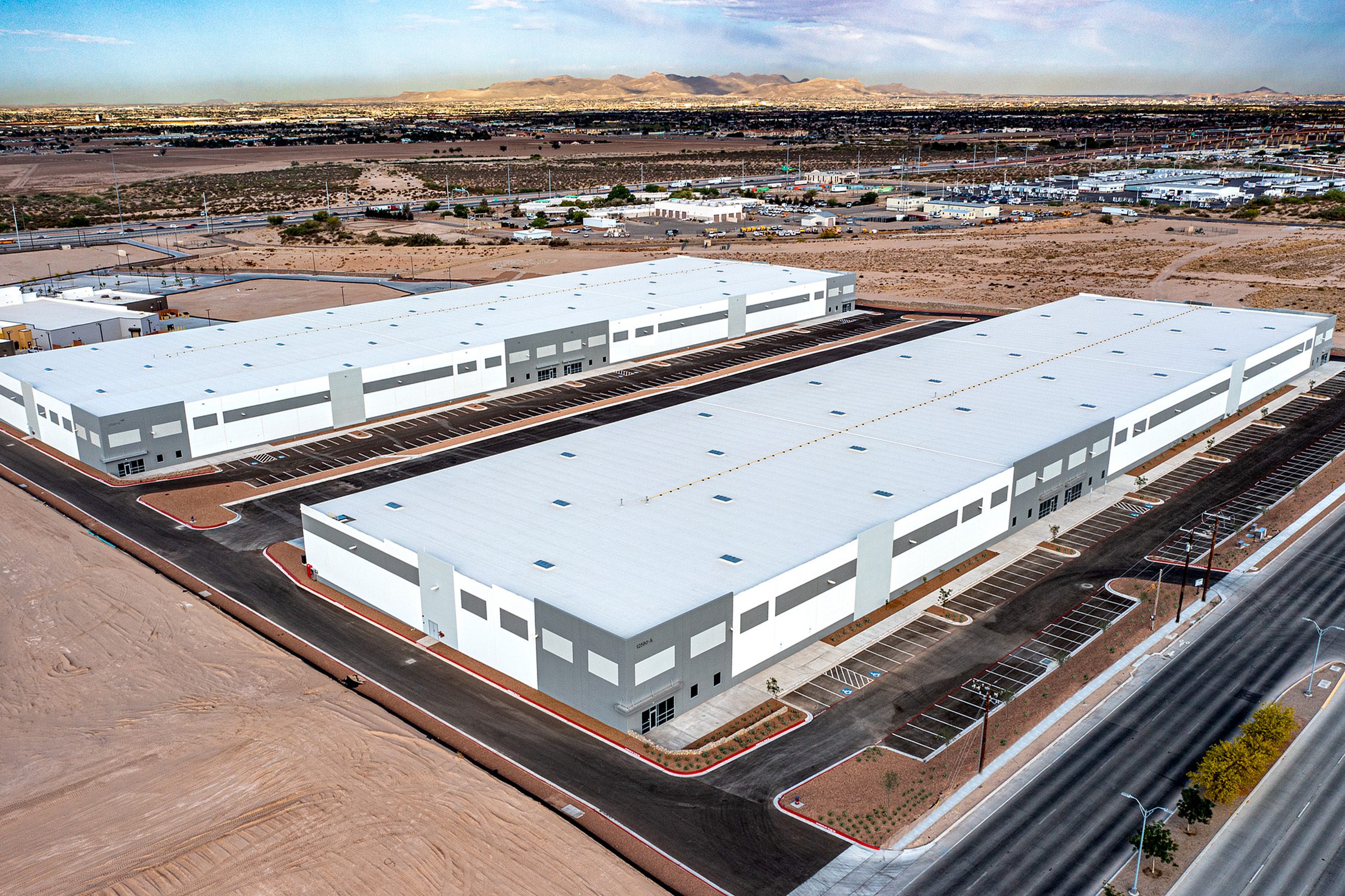 Two large, white and gray industrial warehouse buildings with flat roofs and adjacent parking lots in a desert landscape, surrounded by roads and distant mountains under a clear sky.